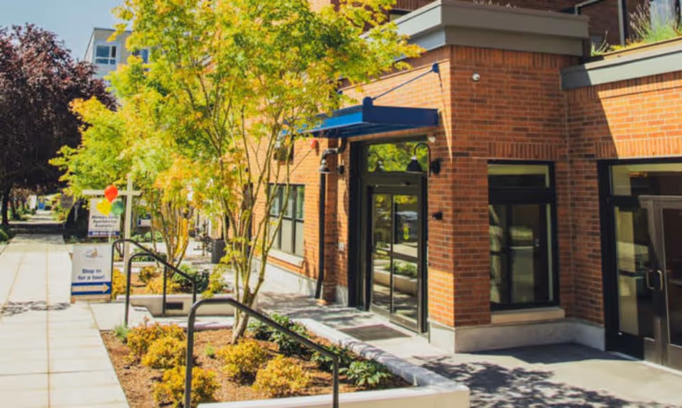 Exterior view of a brick building with large windows and a blue awning over the entrance. There are trees and landscaped plants along the sidewalk, and a sign with balloons that says 'Stop in for a tour!'