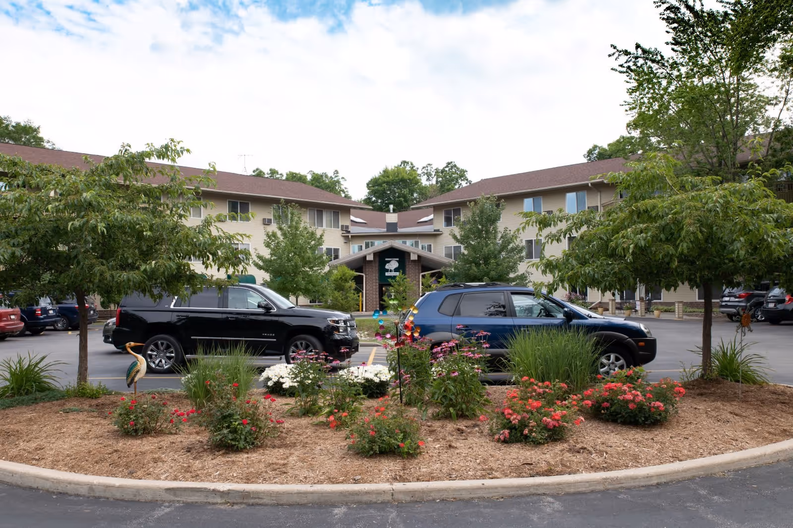 View of the front exterior of The Arboretum Senior Living facility showing a two-story building with beige siding and brown roof. There are trees and flower beds with colorful flowers in the foreground, and several parked cars in the parking lot.