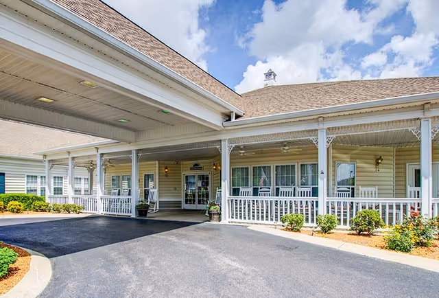 Exterior view of The Bungalows at Mayfield senior living facility entrance with a covered driveway, white railing, rocking chairs on the porch, and a clear blue sky with some clouds.
