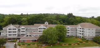Exterior view of a large multi-story senior living facility named The Gables Of Fitchburg, surrounded by greenery and trees under a cloudy sky.