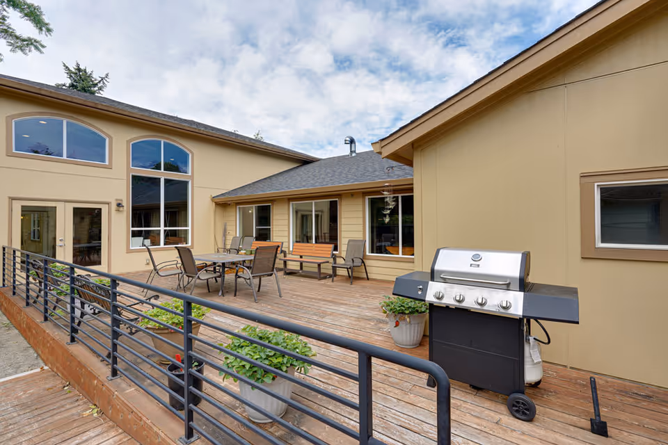 A wooden outdoor deck with a dining table and chairs, bench seating, potted plants, and a gas grill in front of a beige building.