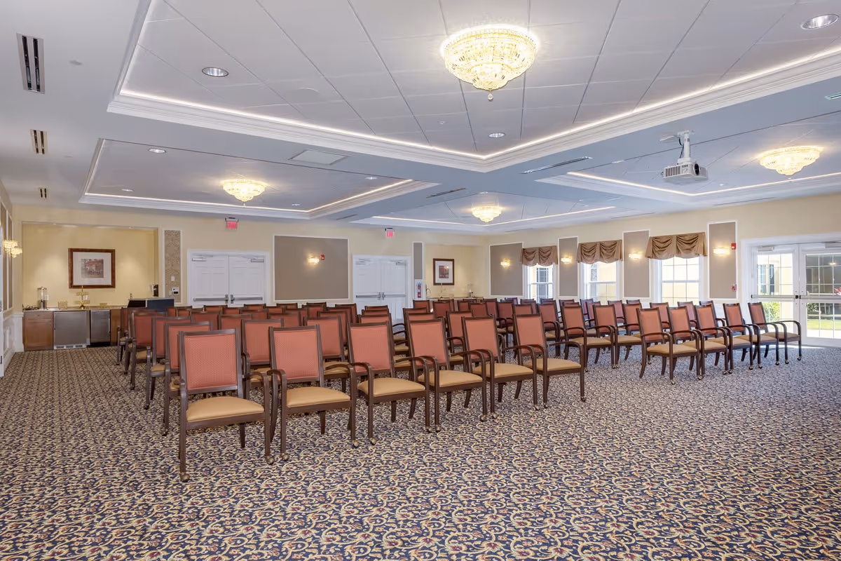 A large, well-lit meeting or event room with rows of wooden chairs featuring red backs and beige seats arranged facing forward. The room has patterned carpet, cream-colored walls with framed artwork, multiple chandeliers on the ceiling, and windows with brown valances letting in natural light. There is a projector mounted on the ceiling and double doors at the back of the room.