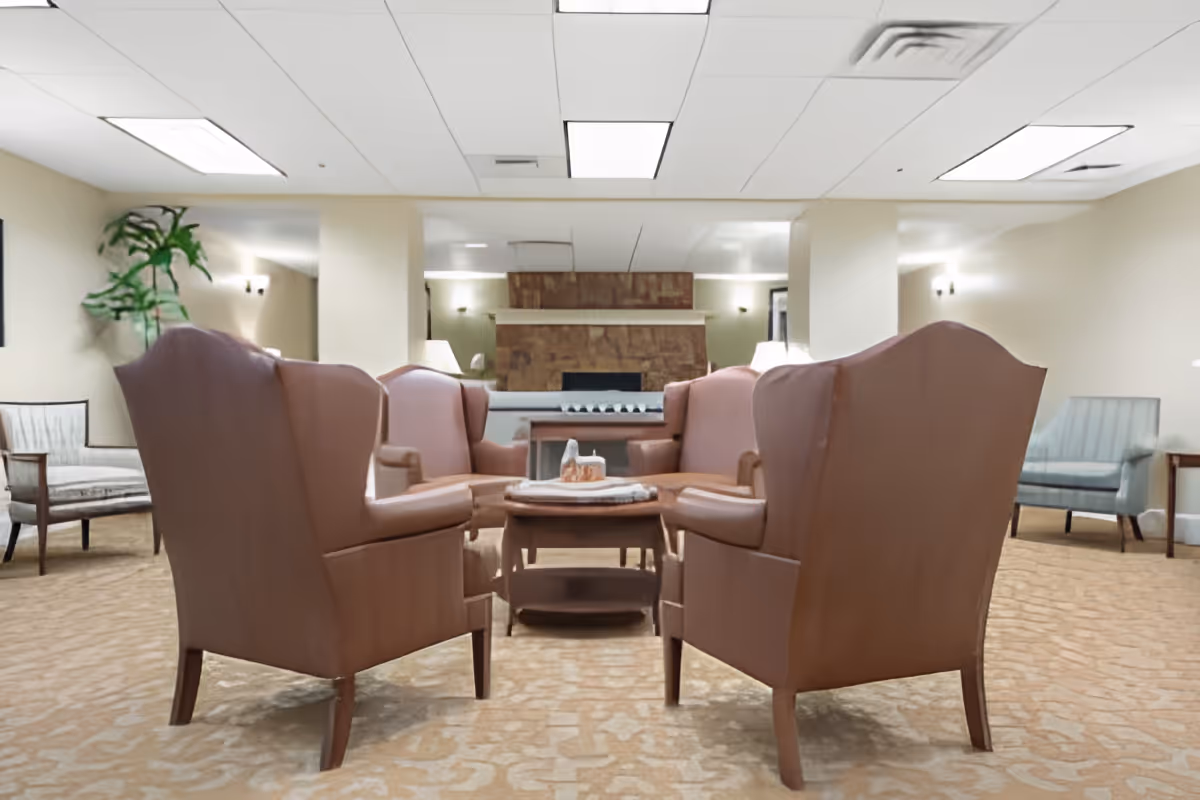 A cozy seating area in a senior living facility with four brown leather armchairs arranged around a wooden coffee table. The room has beige walls, carpeted floor, and a stone fireplace in the background. Additional chairs and lamps are placed along the walls, and a tall green plant is visible in the corner.