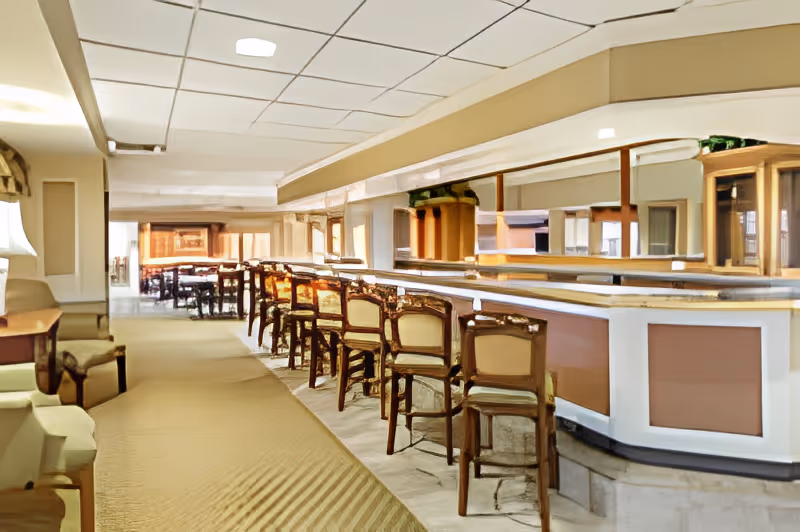 Interior view of a lounge area with a long bar counter and multiple wooden chairs lined up along it. There are cushioned armchairs on the left side and a large mirror behind the bar reflecting the room. The ceiling has recessed lighting and the floor is carpeted.