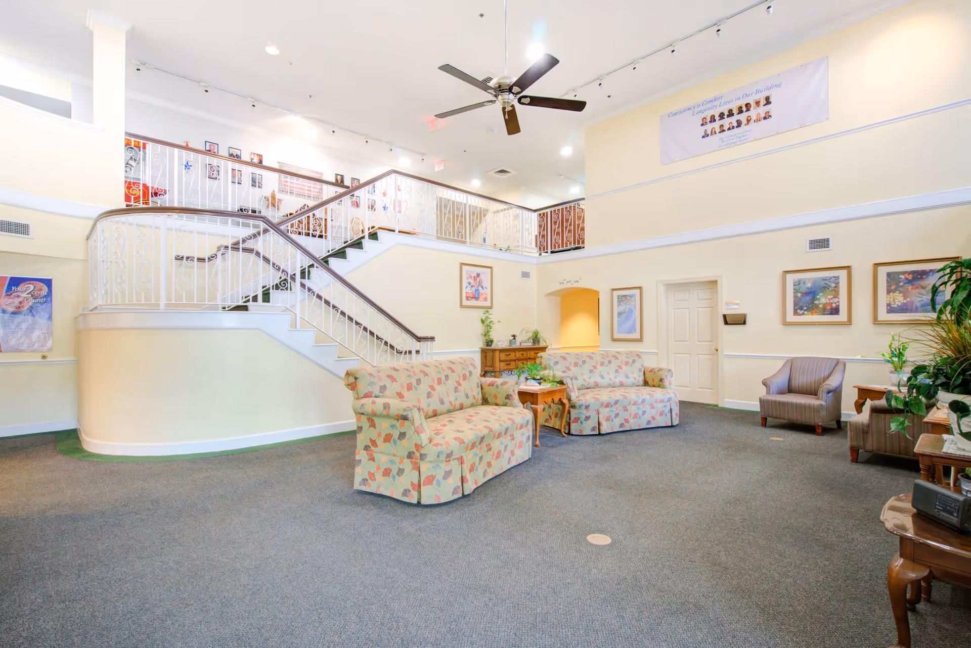 A spacious senior living facility common area with two floral-patterned sofas, two armchairs, a ceiling fan, and a staircase with white railings leading to an upper level. The walls are decorated with framed artwork and a banner, and there are several plants and wooden furniture pieces around the room.