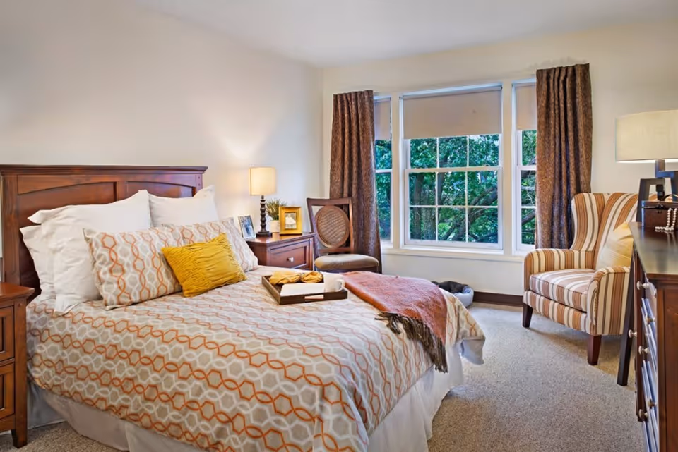 A cozy bedroom with a wooden bed frame, patterned bedspread, multiple pillows including a yellow accent pillow, a wooden nightstand with a lamp and picture frames, a chair near a large window with brown curtains, and a striped armchair next to a wooden dresser with a lamp.