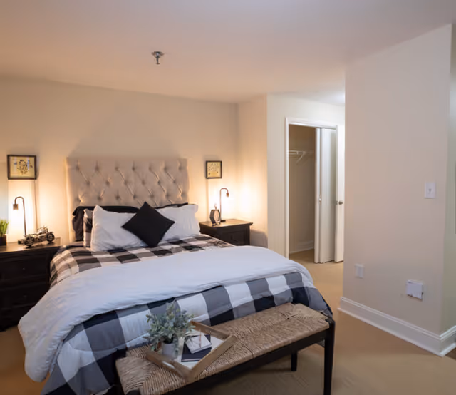 A neatly made bed with a tufted headboard, bedside tables and lamps, and a woven bench at the foot in a neutral-toned bedroom.