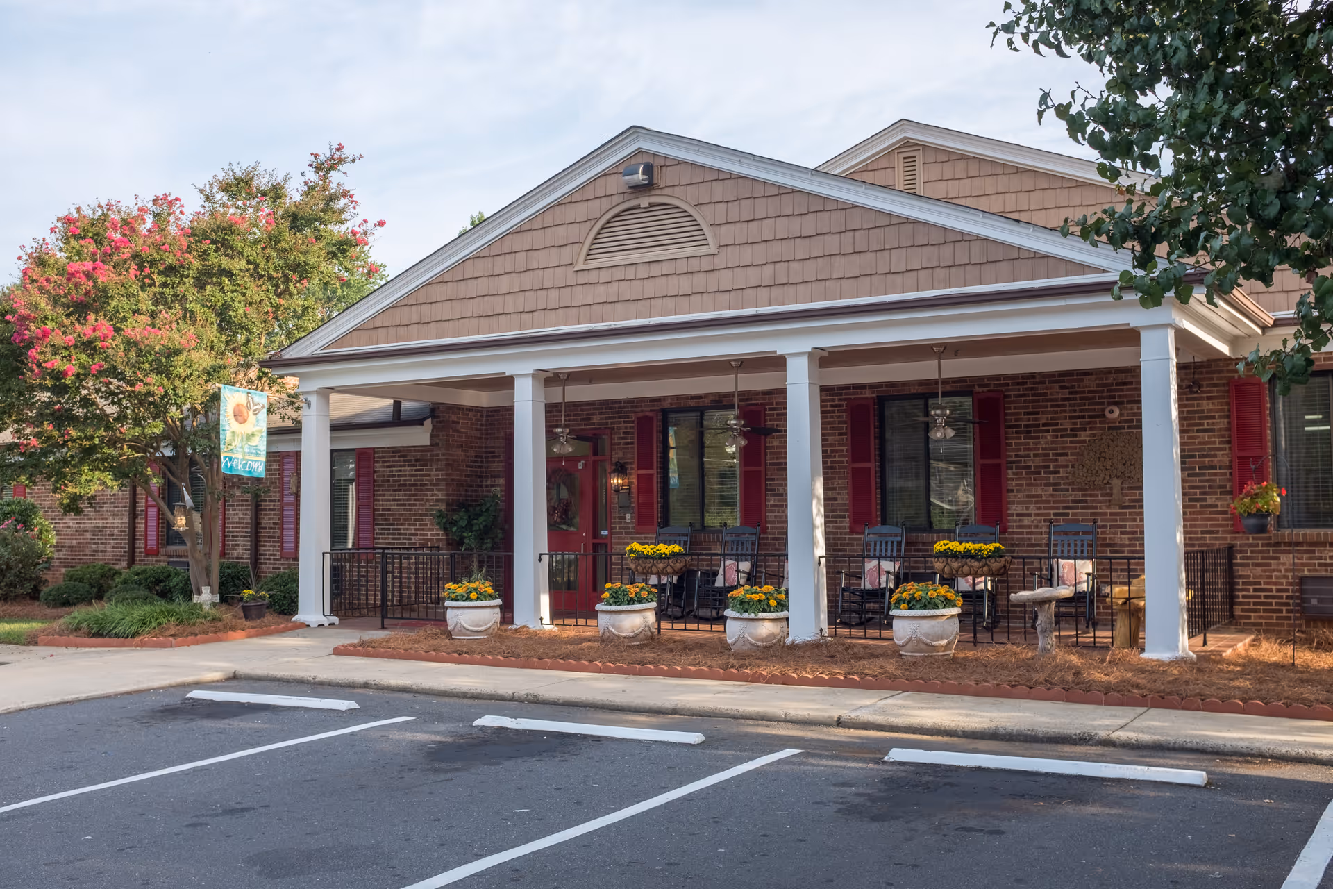 Front entrance of a brick senior living facility with a covered porch, white columns, potted flowers and outdoor chairs.