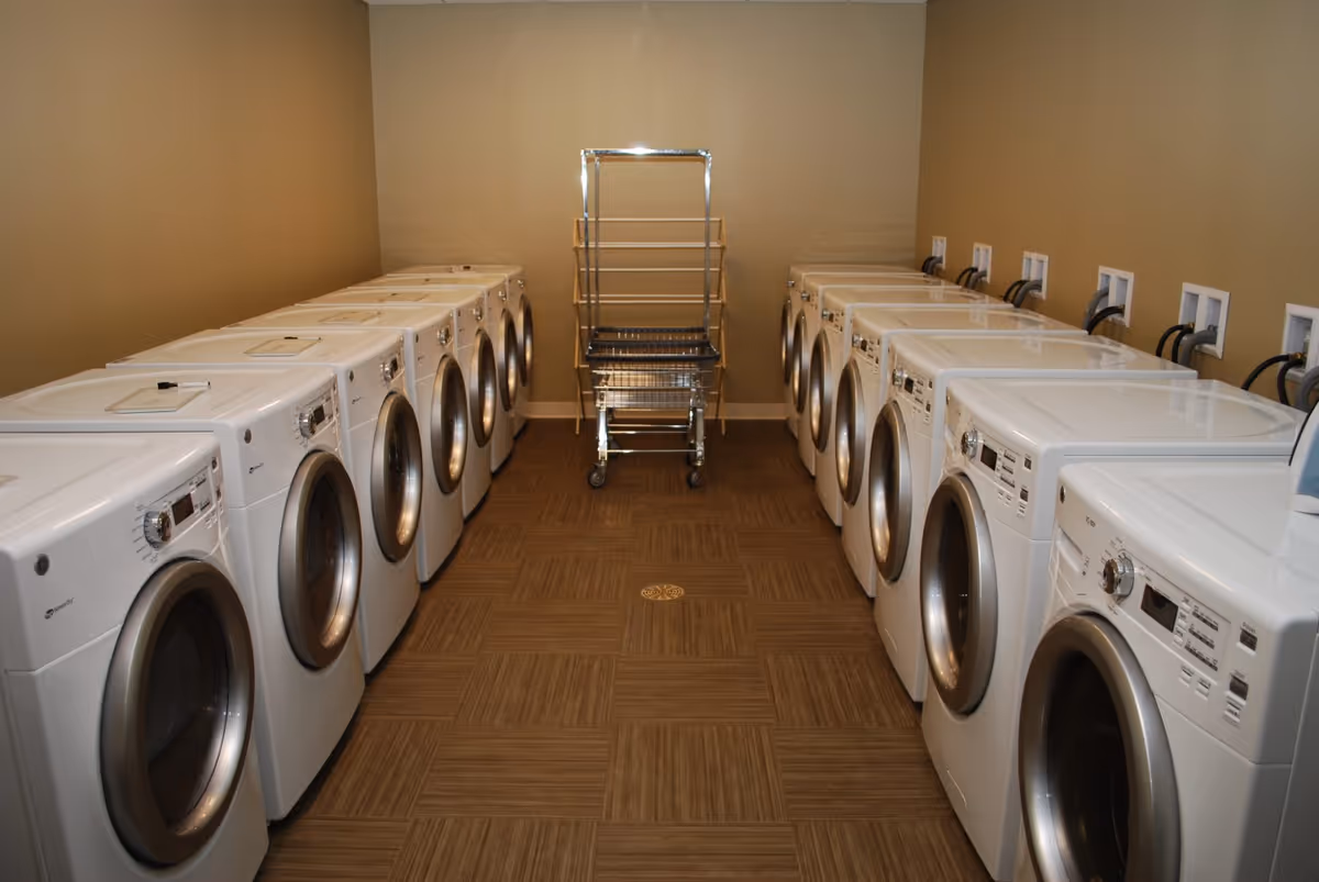 A laundry room with two rows of front-loading washers and dryers facing each other and a laundry cart at the far end.