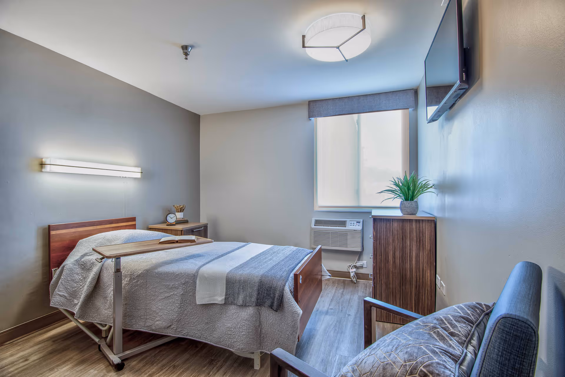 A small, neatly arranged bedroom in a senior living facility with a single bed covered in a white and gray blanket. There is a wooden bedside table with a clock and a small decorative item. A window with a blind lets in natural light, and below it is an air conditioning unit. A wooden cabinet with a potted plant on top is placed against the wall, and a flat-screen TV is mounted above it. A cushioned chair with a patterned pillow is positioned near the bed. The room has light-colored walls and wood flooring.