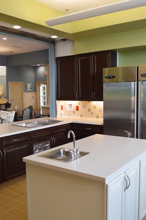 Modern kitchen area with a white island countertop featuring a small sink and faucet. The kitchen has dark wood cabinets, a stainless steel refrigerator, and a tiled backsplash with under-cabinet lighting. In the background, there is a glimpse of a dining or living area with chairs and a table.