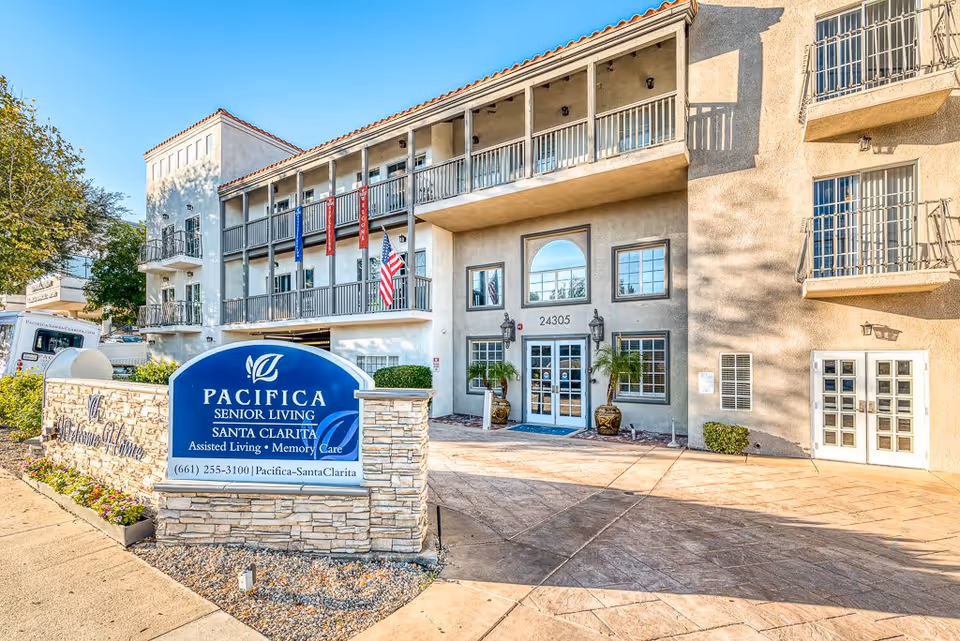 Exterior view of Pacifica Senior Living Santa Clarita building with a stone sign in front displaying the facility name and contact information. The building has balconies, an American flag, and a clear blue sky above.
