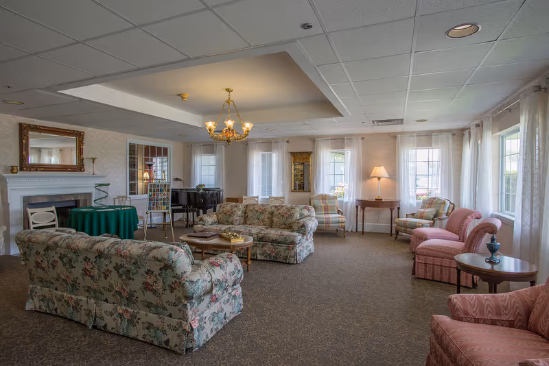 A spacious living room with floral and patterned upholstered sofas and armchairs arranged around a coffee table. The room features large windows with sheer white curtains, a chandelier hanging from a recessed ceiling, a fireplace with a mirror above it, a green-covered round table, a piano, and a floor lamp on a side table.