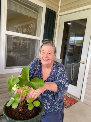 An elderly woman with gray hair tied back is sitting on a porch or patio, smiling and holding a potted strawberry plant with green leaves and small white flowers. Behind her is a beige house exterior with a window, a door with glass panels, and a red doormat.