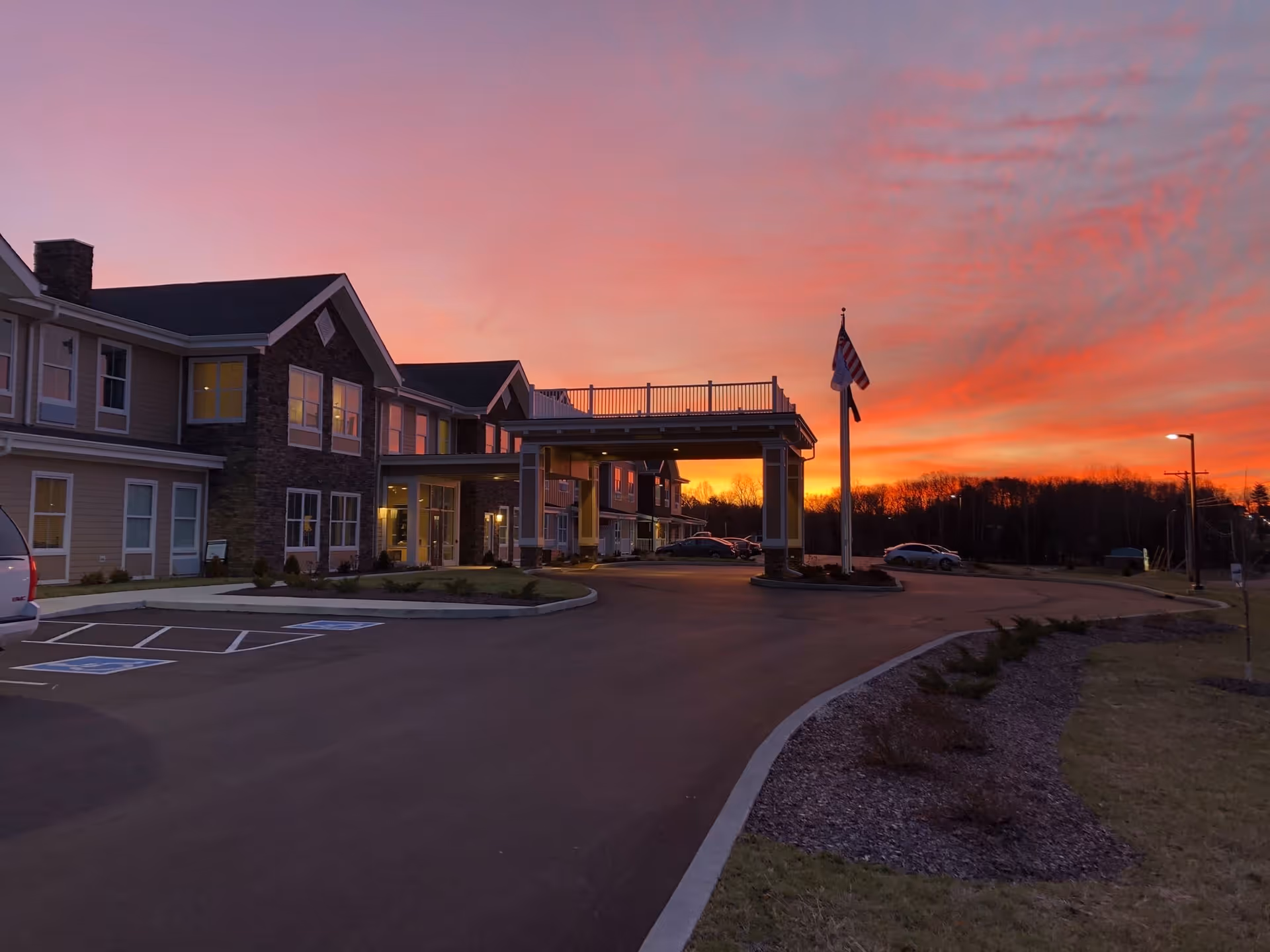 Front entrance and porte-cochère of a senior living building at sunset with a flagpole and parked cars.