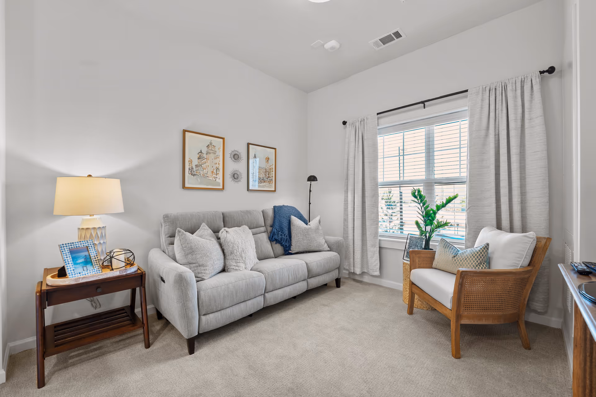 A cozy living room with a light gray sofa adorned with multiple cushions and a blue throw blanket. Next to the sofa is a wooden side table with a lamp, a framed photo, and decorative items. A wooden armchair with white cushions and a patterned pillow is placed near a window with white curtains. The walls are white with two framed pictures and decorative wall hangings. The floor is carpeted in a light beige color.