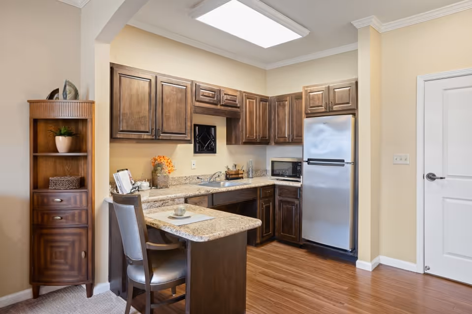 A compact kitchen area with wooden cabinets, a stainless steel refrigerator, a microwave, and a small countertop with a chair. There is a wooden shelving unit with decorative items on the left side and a white door on the right. The floor is wood and the walls are painted beige.