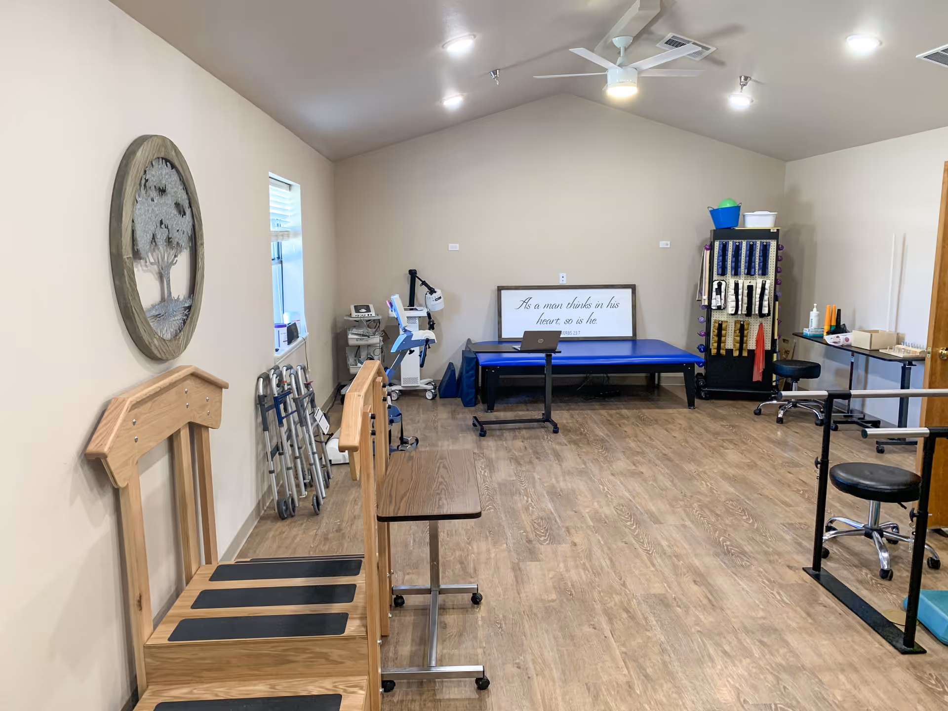 A therapy or rehabilitation room with wooden exercise equipment, walkers, a blue therapy table, a laptop, various exercise bands and equipment on a rack, and a motivational framed quote on the wall. The room has wood flooring, beige walls, and a ceiling fan with lights.