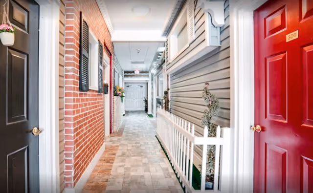 Indoor hallway of a senior living facility with a brick wall on the left and gray siding on the right, featuring several doors including a black door with a hanging flower pot and a red door numbered 306. The hallway has tiled flooring and decorative plants along a white picket fence on the right side.