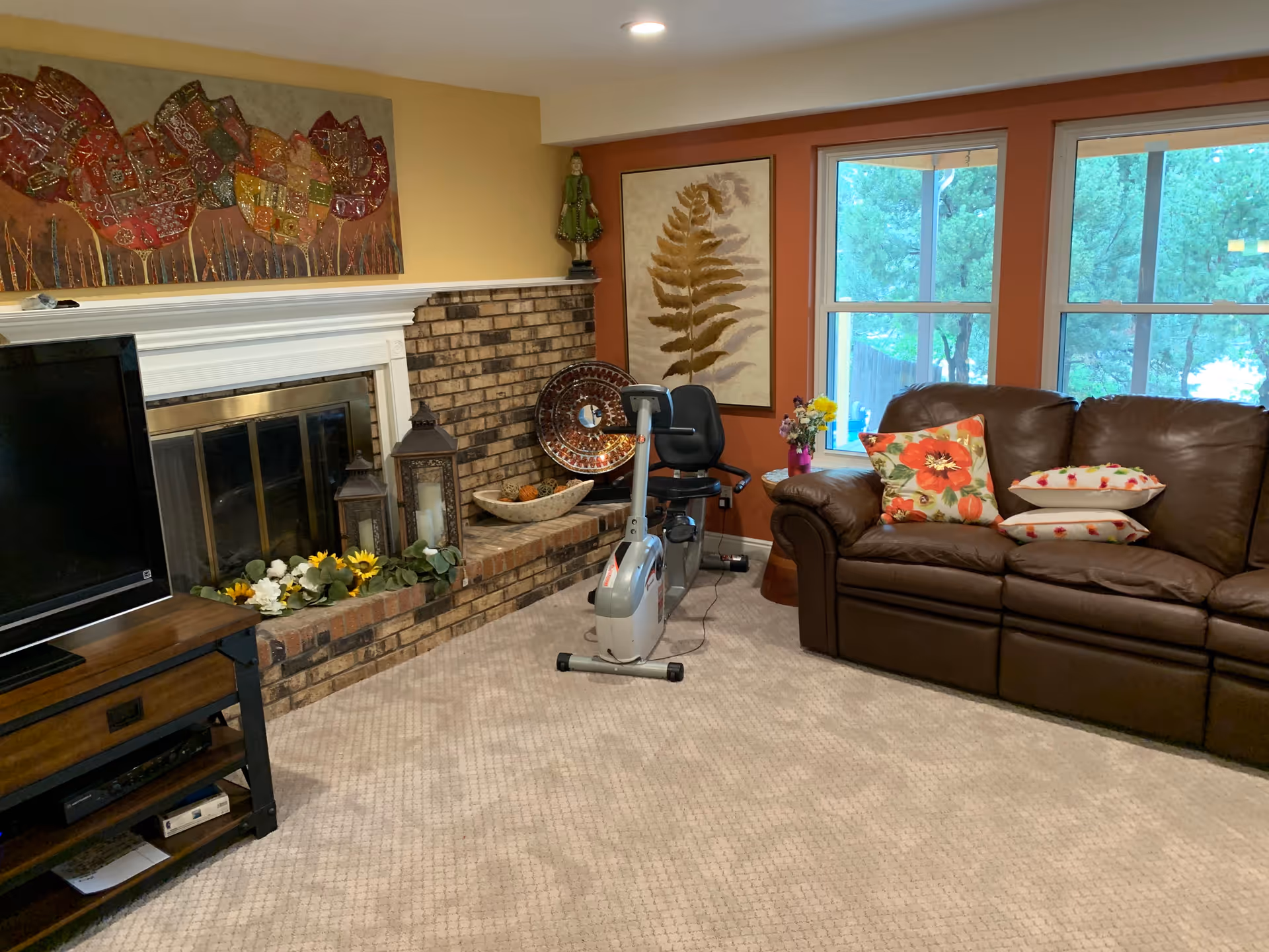 A cozy living room with a brown leather sofa adorned with floral and white pillows, a stationary exercise bike, a brick fireplace decorated with lanterns and flowers, a flat-screen TV on a wooden stand, and two large windows showing green trees outside. The walls are painted in warm tones with framed artwork hanging above the fireplace and near the windows.