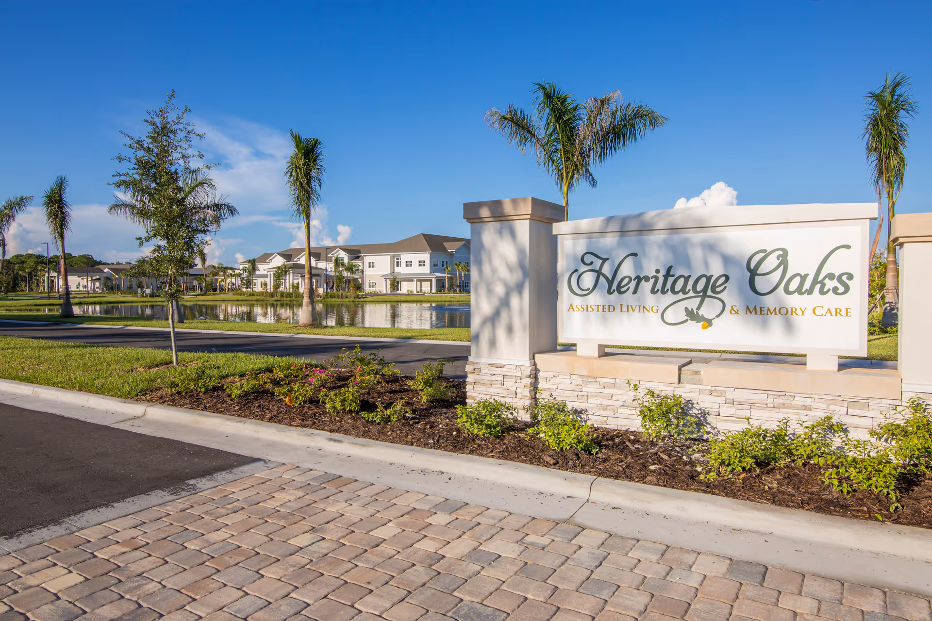 Entrance sign for Heritage Oaks Assisted Living & Memory Care with landscaped greenery, palm trees, a paved road, and a pond with buildings in the background under a clear blue sky.