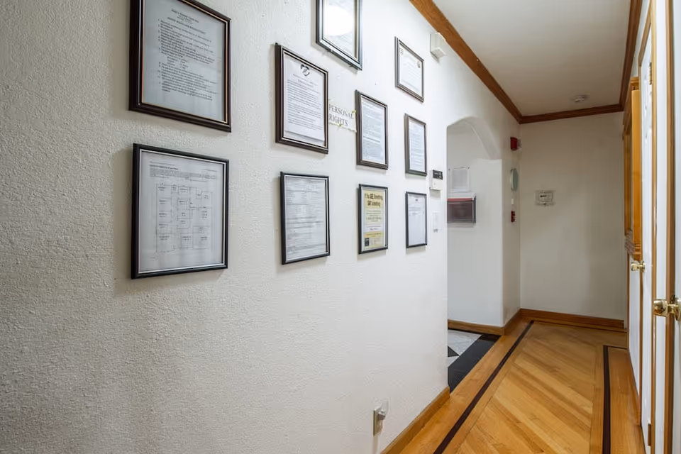 A hallway with wooden flooring and white walls adorned with multiple framed documents and notices. The hallway leads to an arched doorway and has wooden trim along the ceiling and floor edges.