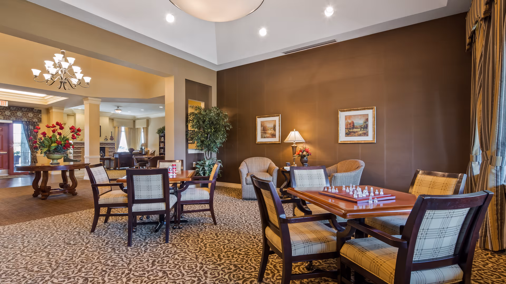 A spacious and well-lit common area in an assisted living facility featuring multiple tables and chairs. One table has a chessboard set up. The room has patterned carpet, beige and brown walls, framed artwork, a potted plant, and large windows with curtains allowing natural light. In the background, there is a seating area with armchairs and a fireplace.