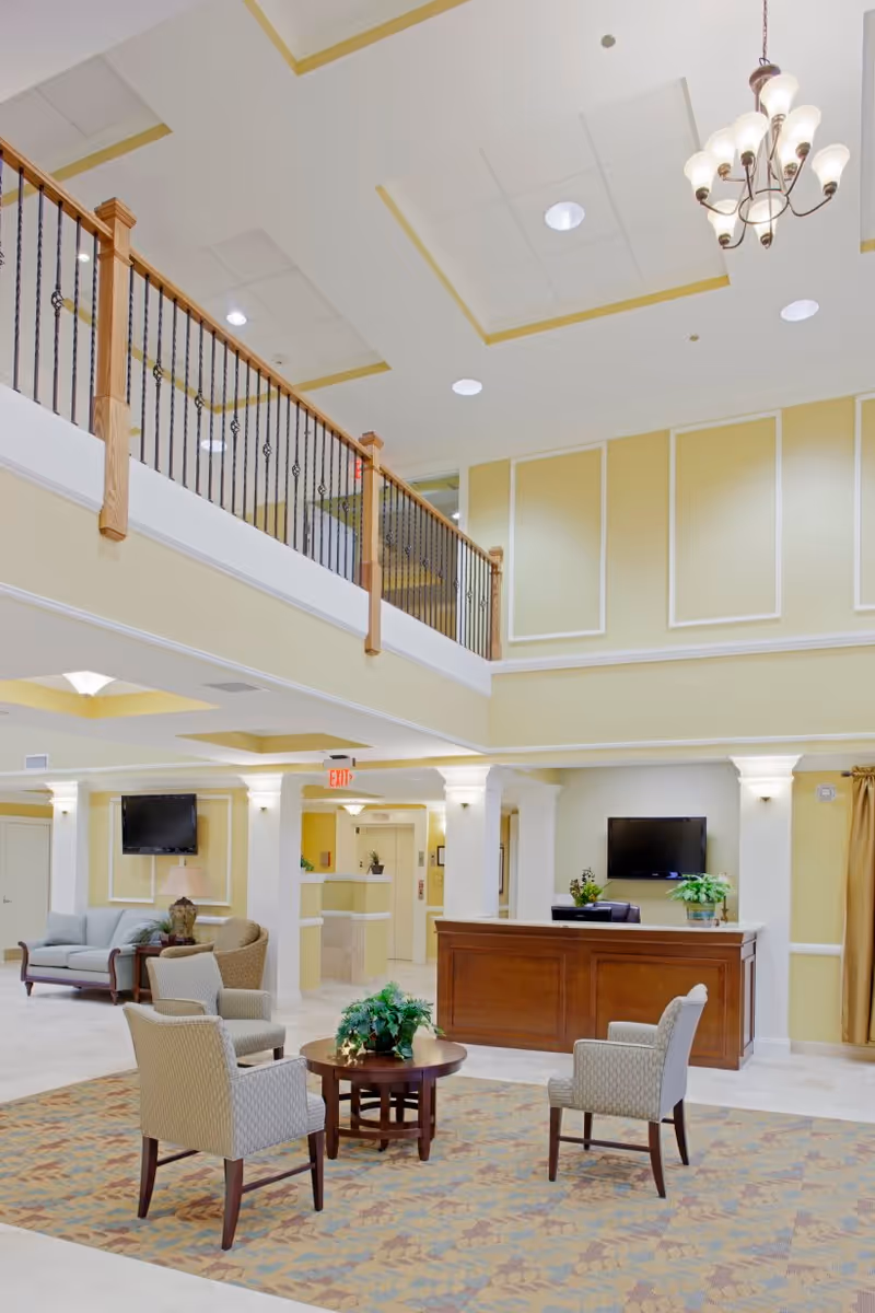 Spacious and well-lit senior living facility lobby with high ceilings, a chandelier, a wooden reception desk, a seating area with four upholstered chairs around a round wooden table with a plant, and a sofa with a side table and lamp. The walls are painted yellow with white trim, and there is a balcony with a wooden railing above.