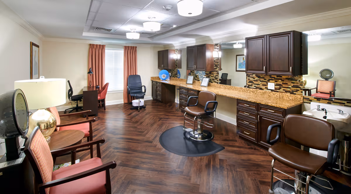 Interior view of a salon area in a senior living facility with two brown salon chairs in front of a long counter with granite top and dark wooden cabinets. The room has wood-patterned flooring, a large mirror, a sink, and several chairs along the walls. There is a window with orange curtains and a desk with a chair in the background.