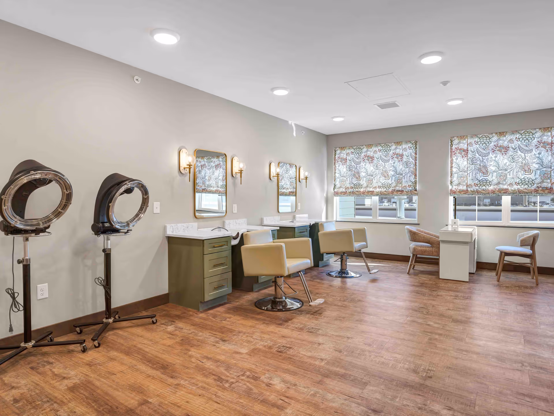 A bright and spacious salon area in Cedarhurst Senior Living of Wentzville featuring two salon chairs in front of mirrors with wall-mounted lights, two hair drying stations on the left, and a small seating area with two chairs and a table near windows with floral-patterned blinds.
