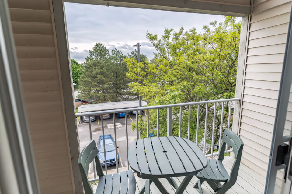 Small balcony with a gray wooden table and two matching chairs. The balcony overlooks a parking lot with several cars and is surrounded by green leafy trees under a cloudy sky.