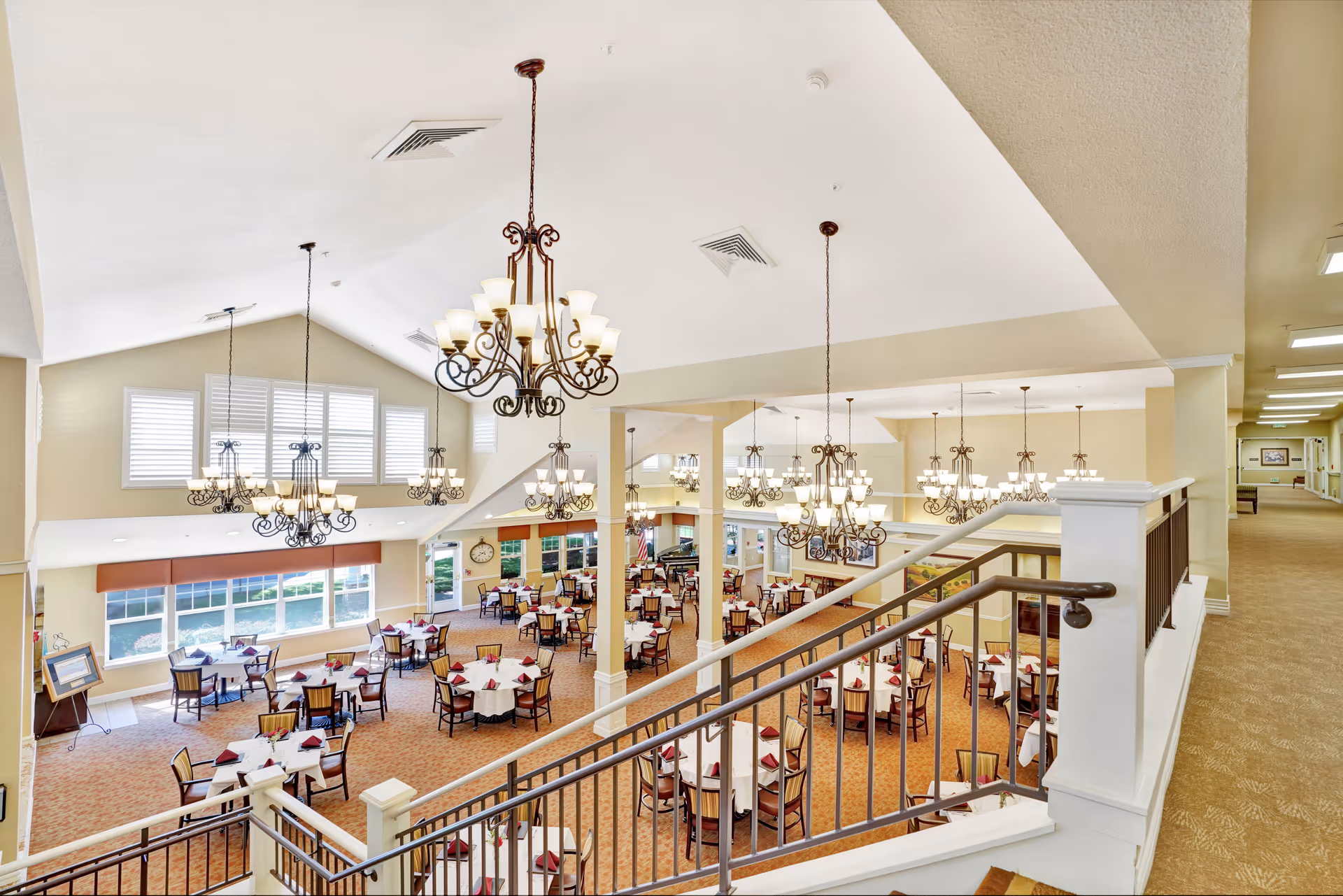 Spacious two-story dining room with multiple round tables set for meals, ornate chandeliers, and a staircase overlooking the area.