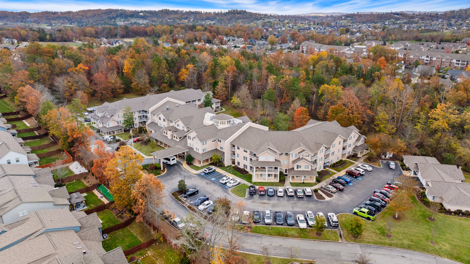 Aerial view of Creekside at Shallowford, a senior living community surrounded by trees with autumn foliage. The multi-story beige buildings have pitched roofs and are arranged around a parking lot filled with cars. The community is situated near a residential neighborhood with hills in the background under a partly cloudy sky.
