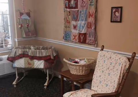 A cozy interior corner of a senior facility with a bassinet, rocking chair, and quilted decor.
