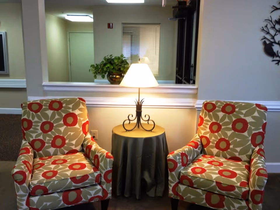 Two armchairs with a floral pattern in red and beige are placed on either side of a small round table covered with a dark cloth. A table lamp with a white shade is on the table, illuminating the area. Behind the table is a half wall with a potted plant and a window with blinds. The walls are light-colored and there is a decorative metal wall art piece on the right.
