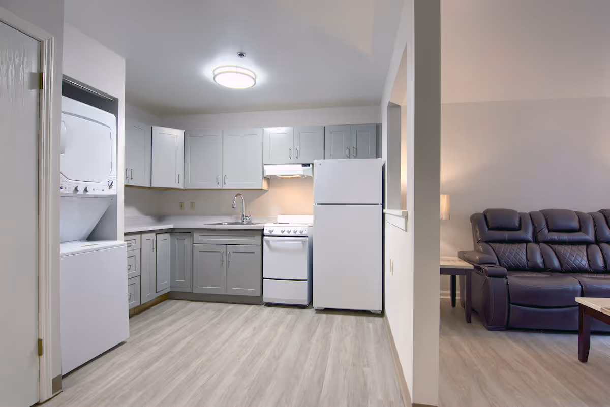 A modern kitchen area with light gray cabinets, a white refrigerator, a white stove with an overhead vent, and a sink with a faucet. To the left, there is a stacked washer and dryer unit. The kitchen opens into a living room area with a dark leather recliner sofa and a side table with a lamp. The flooring is light wood, and the walls are painted white.