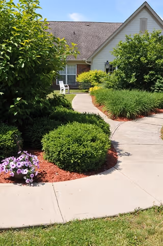 A curved concrete walkway leading to the entrance of a house with a sloped roof. The walkway is bordered by well-maintained green bushes, flowering plants, and red mulch. There is a white chair near the house entrance and a clear blue sky above.