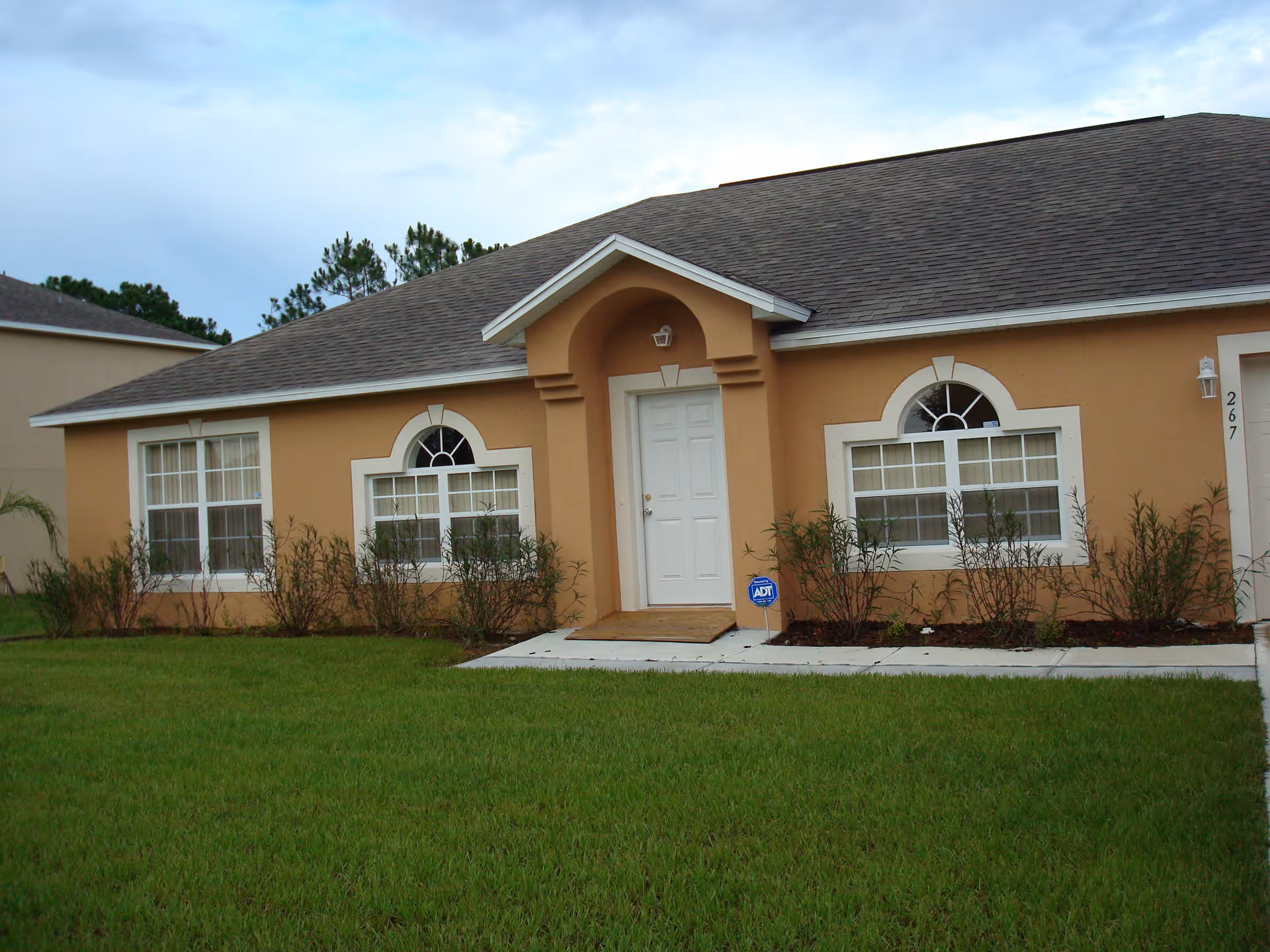 Exterior view of a single-story house with a tan facade, white trim around windows and door, a gray shingled roof, and a green lawn in front. There are three windows visible, two with arched tops, and a white front door under a small covered porch. Some small shrubs are planted along the front of the house.