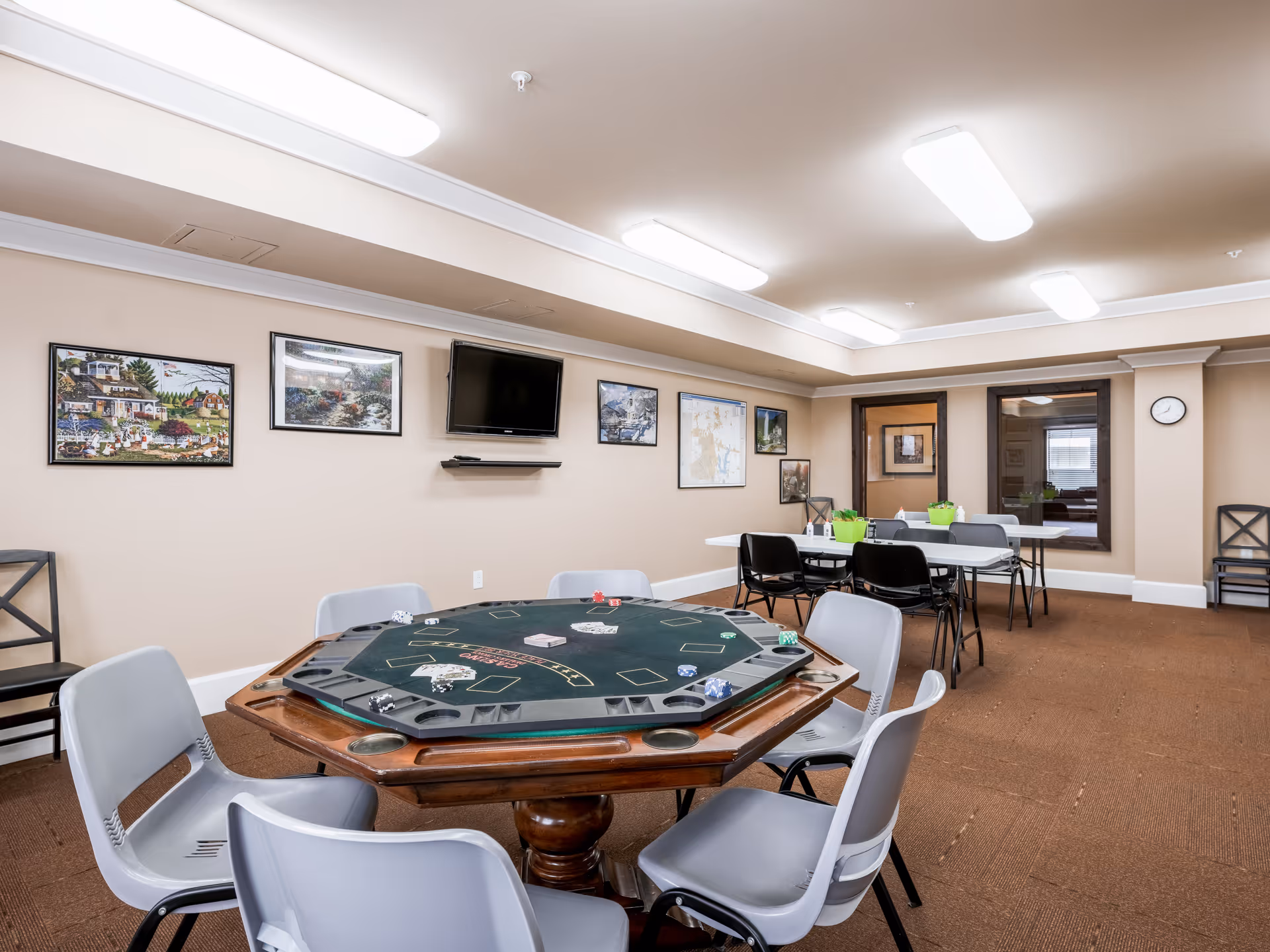 A well-lit game room with a large octagonal poker table surrounded by gray chairs in the foreground. In the background, there are two rectangular tables with black chairs and green potted plants on top. The beige walls are decorated with framed pictures and a mounted flat-screen TV. The room has a brown carpet and fluorescent ceiling lights.