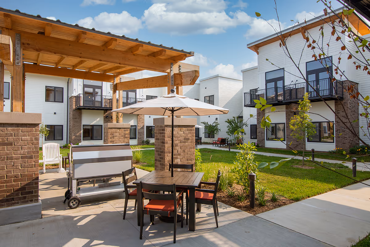 Outdoor courtyard area at Preston Greens Senior Living featuring a wooden pergola, a table with four chairs and a large umbrella, a grill, green lawn, young trees, and modern two-story white buildings with balconies in the background under a partly cloudy sky.