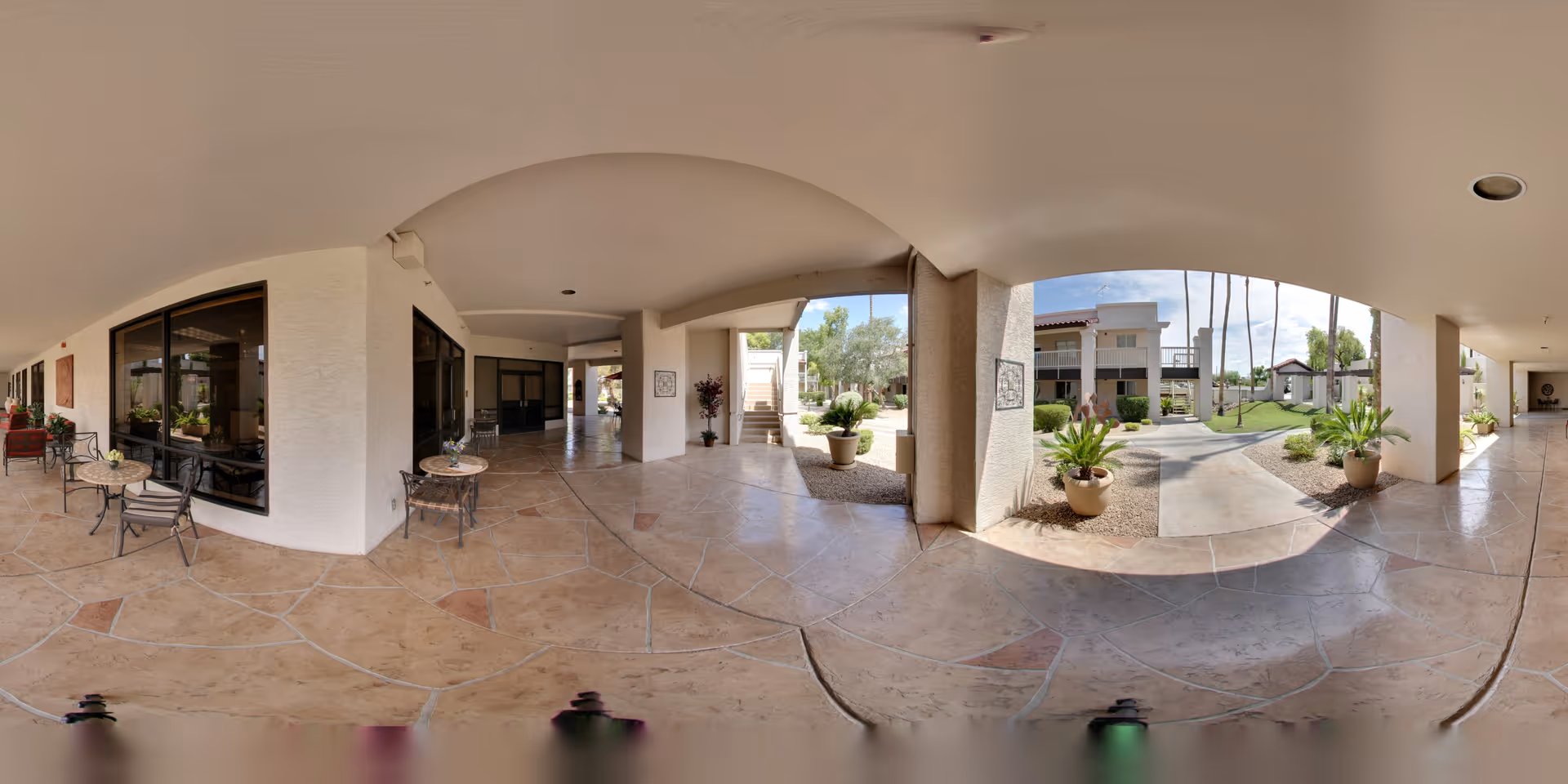 Covered outdoor corridor and patio with seating, potted plants, and archways opening to a landscaped courtyard and adjacent senior living buildings.