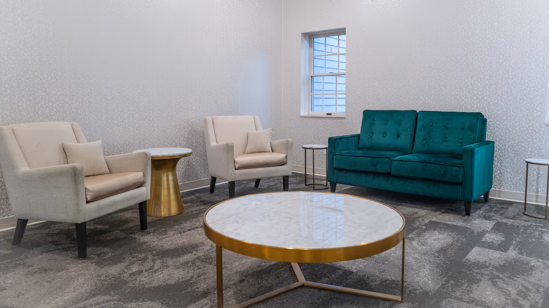 A small sitting area with two beige armchairs, a teal loveseat, a round marble coffee table with a gold base, and two small round side tables. The room has a window with white trim and light-colored patterned wallpaper.
