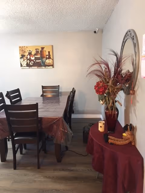 A dining area with a rectangular table covered with a plastic tablecloth and surrounded by six wooden chairs. On the right side, there is a small round table draped with a burgundy cloth, holding a vase with red flowers and decorative grasses, a candle, and a small stuffed dog. A round mirror hangs on the wall above the small table, and a painting of pottery and flowers is on the far wall.