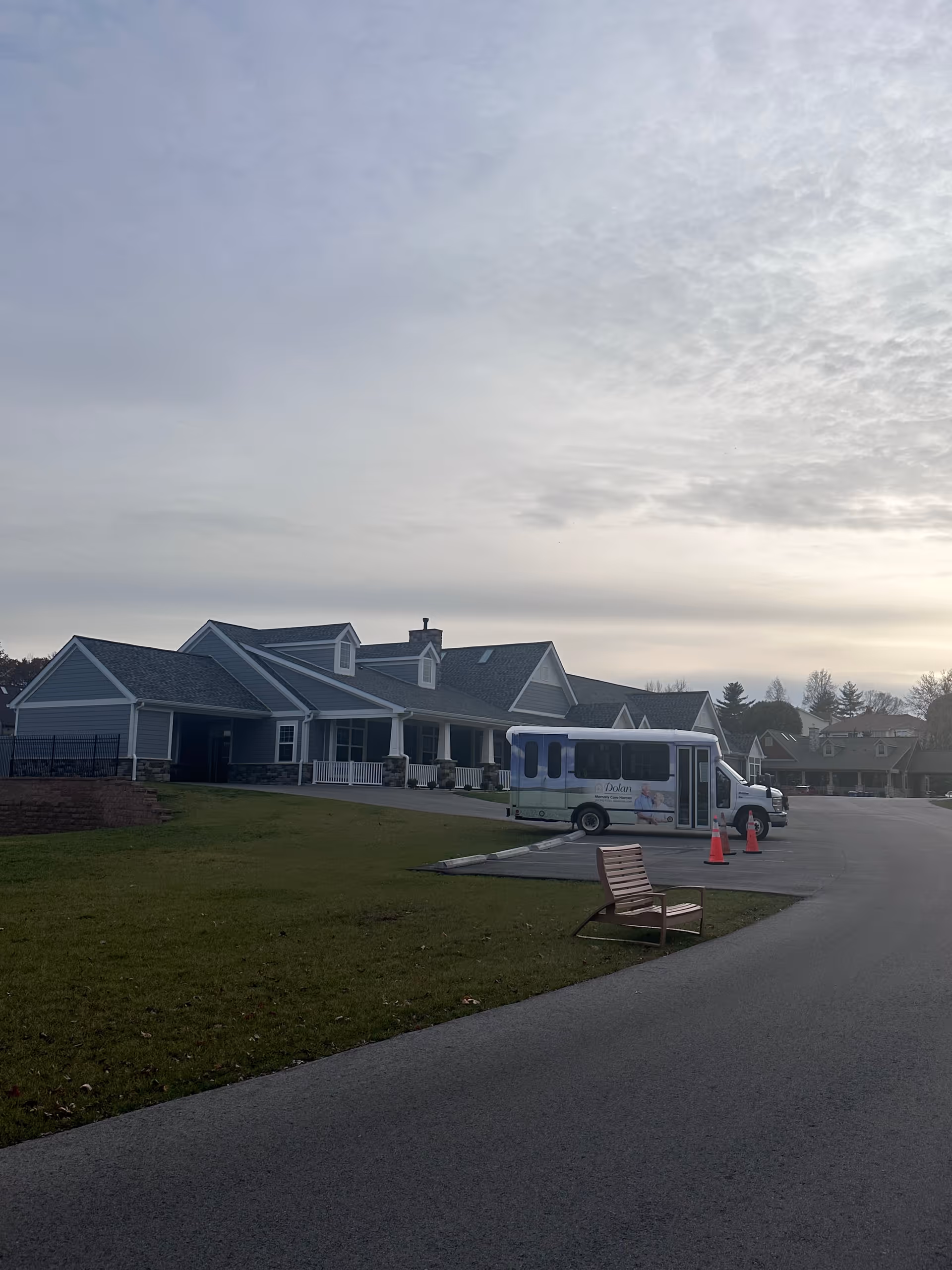 Exterior view of a single-story building with a gray roof and blue siding, a small parking lot with a shuttle bus and orange traffic cones, a wooden bench on the grass near the road, and a cloudy sky overhead.