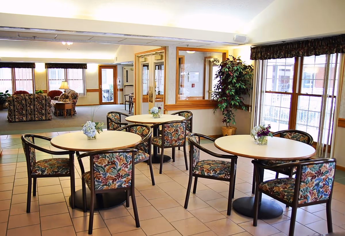 A bright, clean common area in a senior living facility featuring round tables with floral-patterned chairs. Each table has a small floral centerpiece. In the background, there is a seating area with floral upholstered sofas and armchairs, a wooden side table with a lamp, large windows with valances, and a potted plant near a set of double doors.