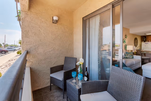 A small outdoor balcony with two wicker chairs and a glass-top table holding a bottle of wine, two blue wine glasses, and a small vase with white flowers. The balcony overlooks a parking lot and is adjacent to a sliding glass door leading into a living room with a white sofa and kitchen area in the background.