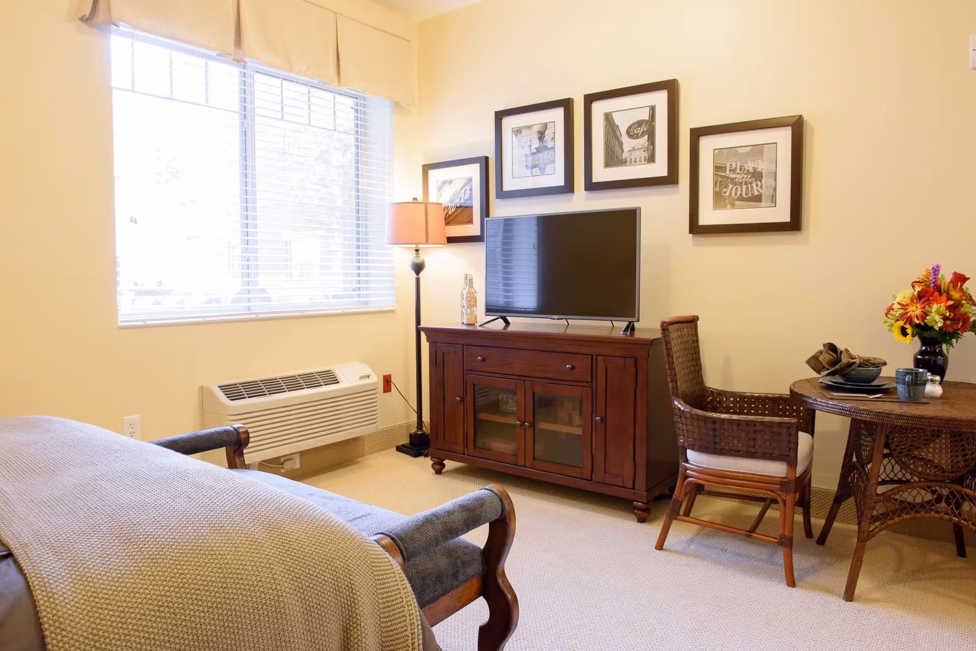 A cozy living room area with a wooden TV stand holding a flat-screen television, a floor lamp, and framed pictures on the wall above. There is a wicker chair and a round wicker table set with plates, cups, and a vase of colorful flowers. A window with blinds lets in natural light, and a cushioned bench or sofa with a knitted throw blanket is partially visible in the foreground.