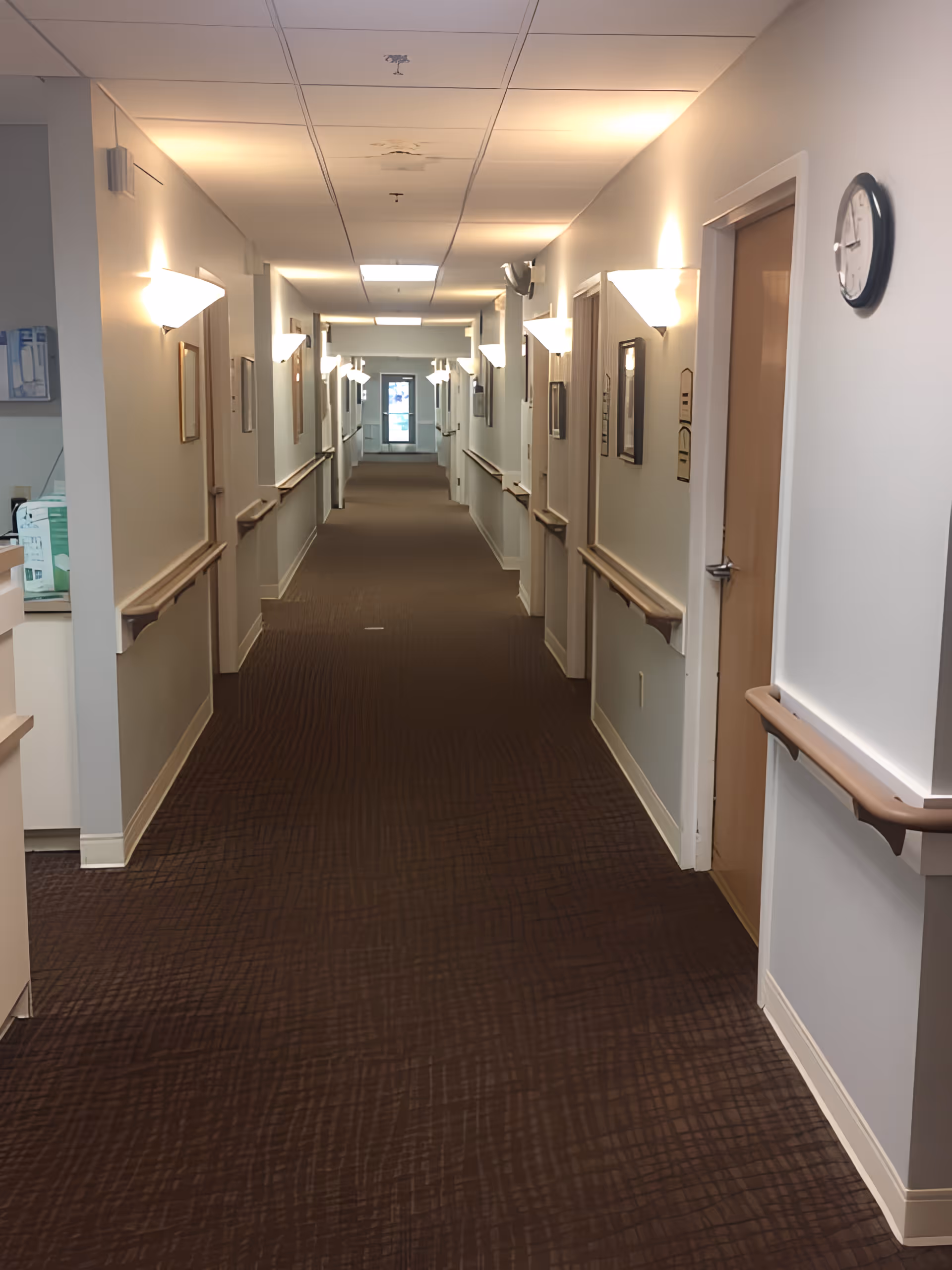 A long, well-lit hallway in a senior living facility with brown patterned carpet and beige walls. There are handrails on both sides of the hallway, several closed wooden doors, wall-mounted lights, framed pictures, and a clock on the right wall. At the end of the hallway, there is a glass door letting in natural light.