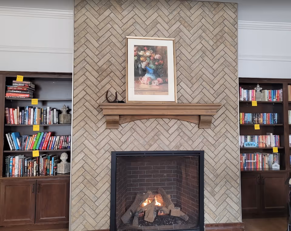 A cozy indoor fireplace with a wooden mantel and a framed painting of flowers above it. On either side of the fireplace are built-in dark wood bookshelves filled with various books, some labeled with yellow sticky notes.