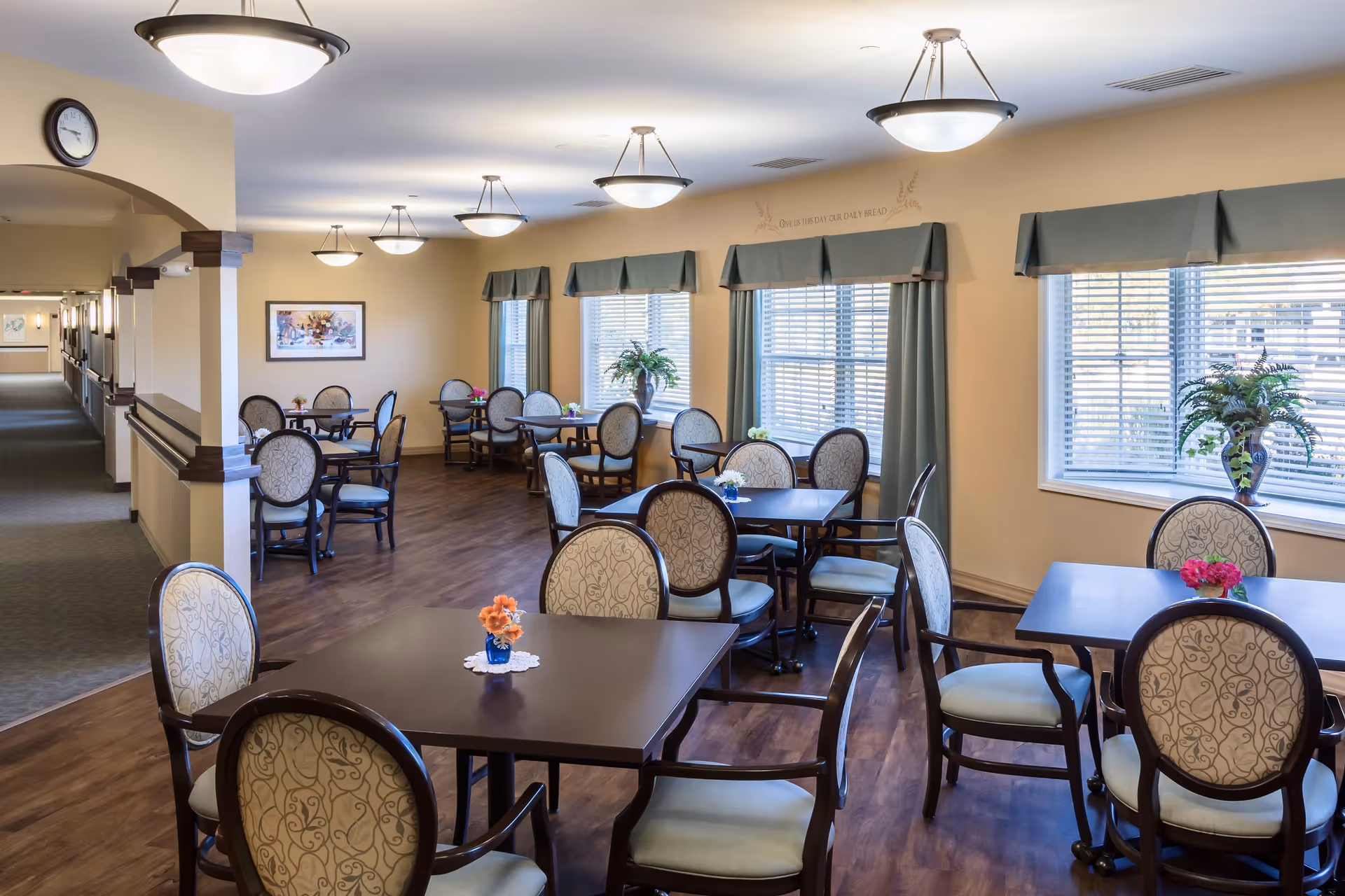 Bright communal dining room with multiple tables and upholstered chairs, pendant lights, and windows with green valances.